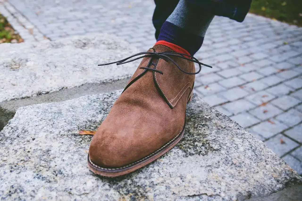 Zapato de serraje marrón con cordones negros, calcetín a rayas y un pie apoyado en una roca. La piel de serraje se puede mojar sin problema.