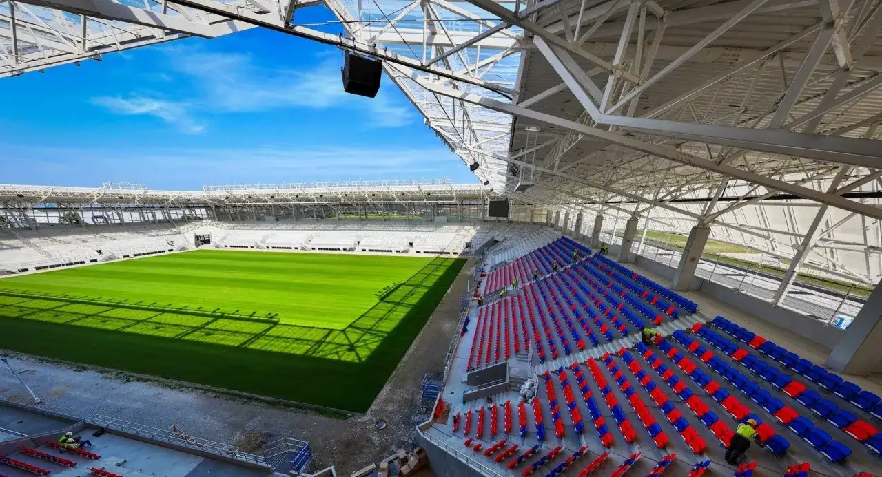Nowy stadion Odra Opole z zieloną murawą i rzędami czerwonych i niebieskich krzesełek pod błękitnym niebem.