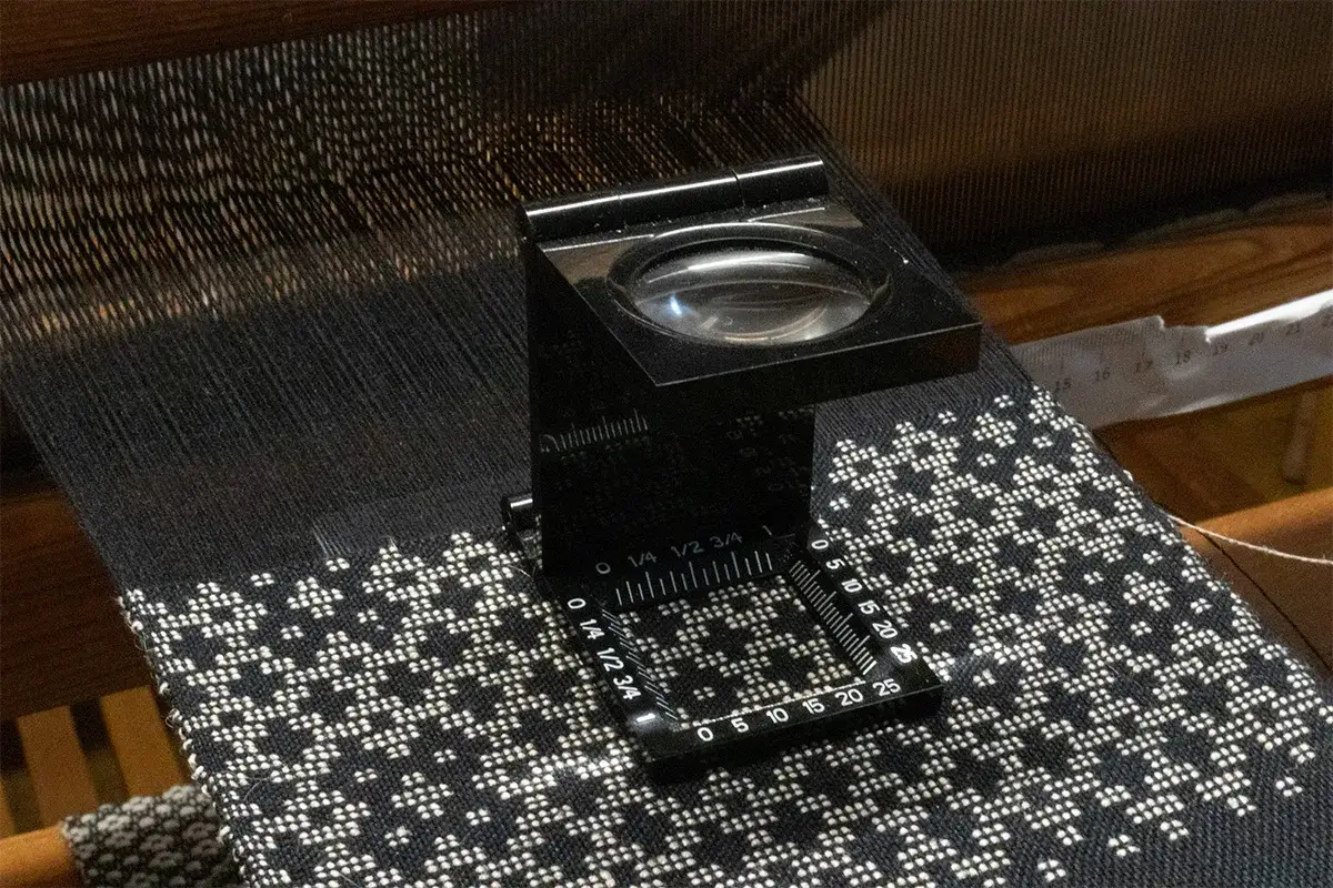 A magnifying tool with a ruler is placed on black fabric with a white pattern. It shows how thread count is measured by counting threads within a square inch.