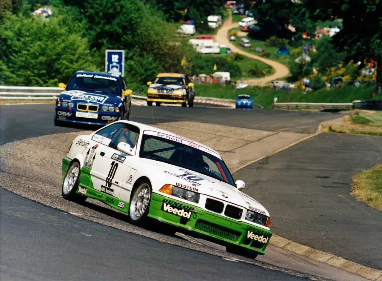 Rennwagen-Action: Ein weißer BMW E36 Motorsport-Bolide mit grünen Akzenten auf der Rennstrecke, verfolgt von anderen Fahrzeugen.