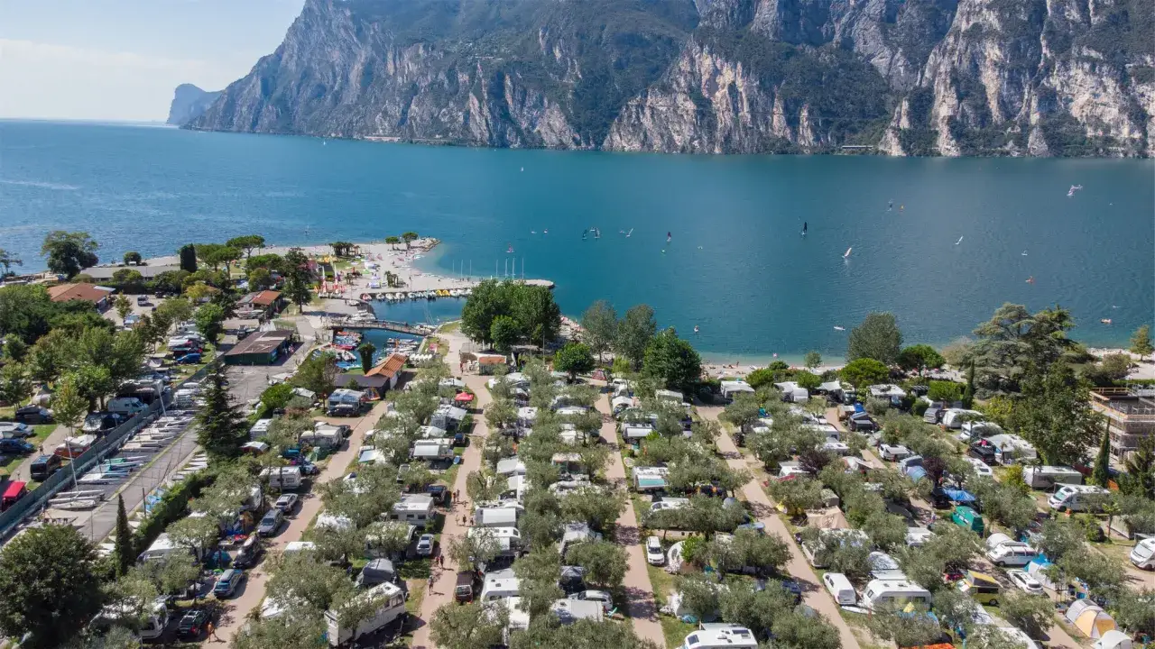 Widok z lotu ptaka na camping Maroadi nad jeziorem, z rzędami kamperów i namiotów, plażą i górami w tle.