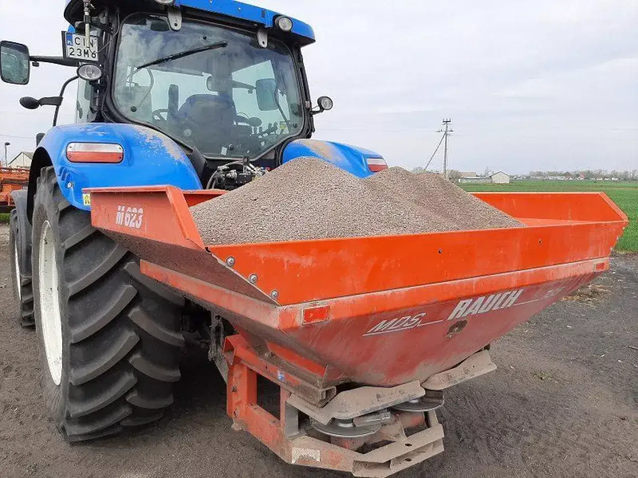 Niebieski traktor z przyczepionym rozsiewaczem napełnionym wapnem dolomitowym, gotowy do pracy na polu.