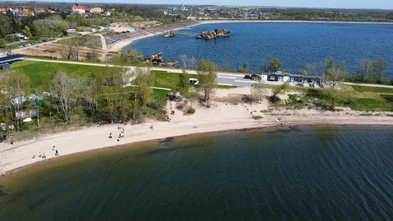 Słoneczna mietków plaża nad jeziorem, z ludźmi spacerującymi po piasku i odpoczywającymi na kocach. W tle widać zabudowania i infrastrukturę.