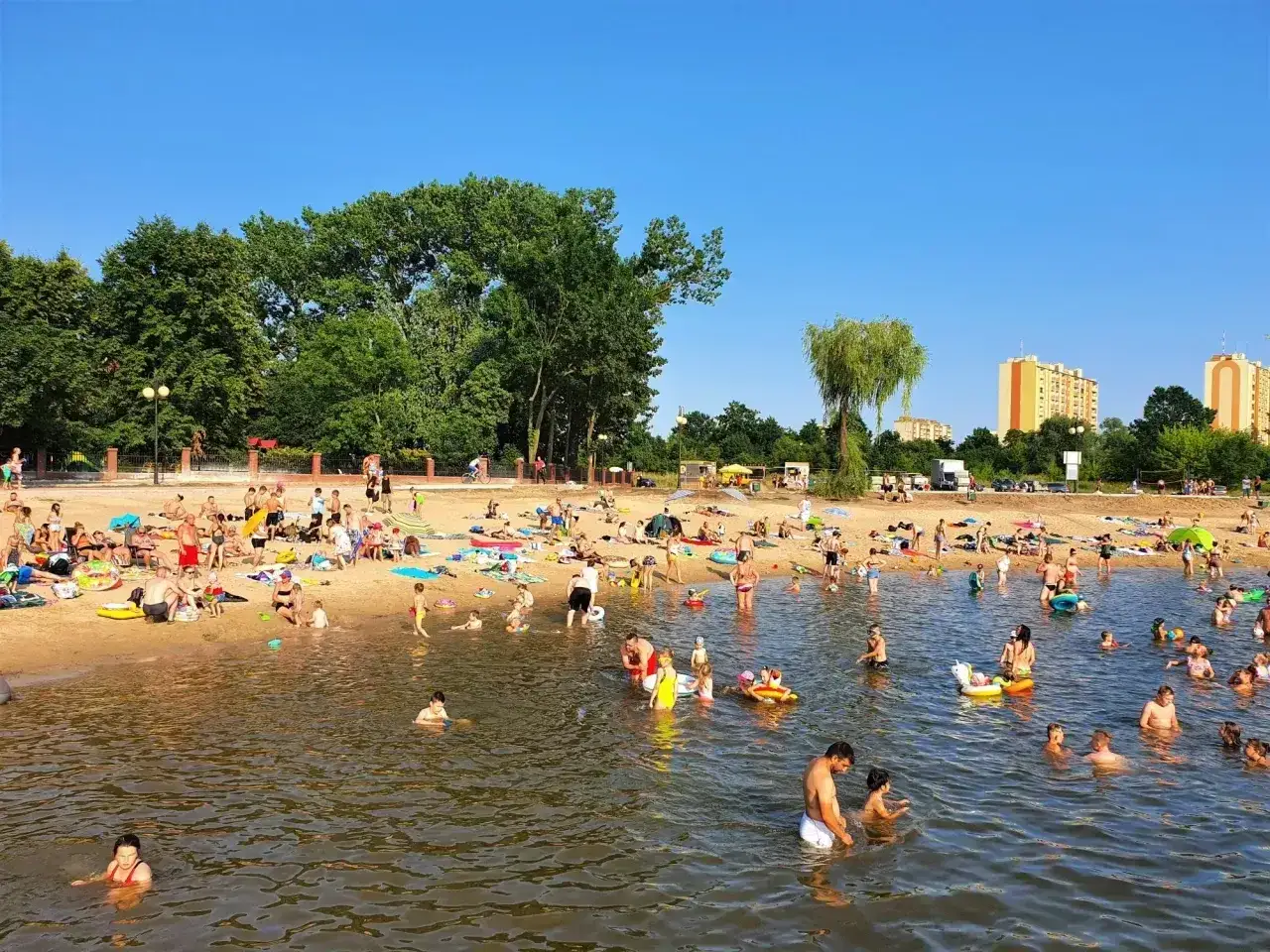 Tłum ludzi odpoczywa na piaszczystej plaży i kąpie się w wodzie. Słoneczny dzień nad zalewem Borki.