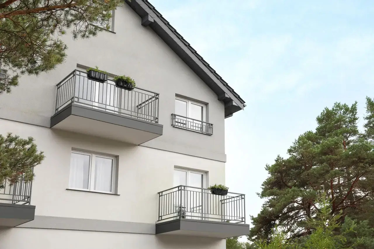 Nowoczesny rodzaj balkonu z metalową balustradą i donicami z zielenią, przylegający do jasnoszarego budynku mieszkalnego.