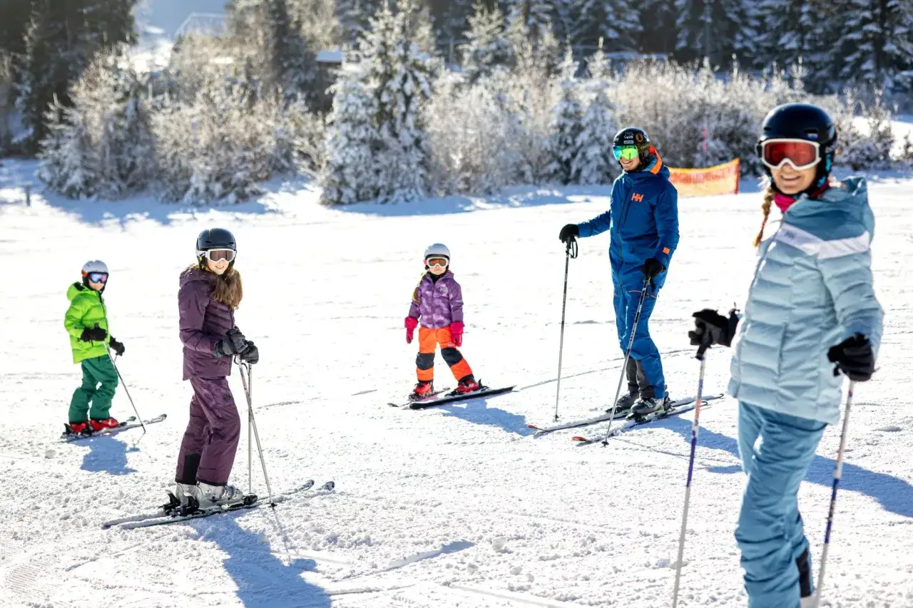 Familie genie&szlig;t einen sonnigen Tag im Lipno Skigebiet. Kinder und Erwachsene auf Skiern, umgeben von verschneiten B&auml;umen.