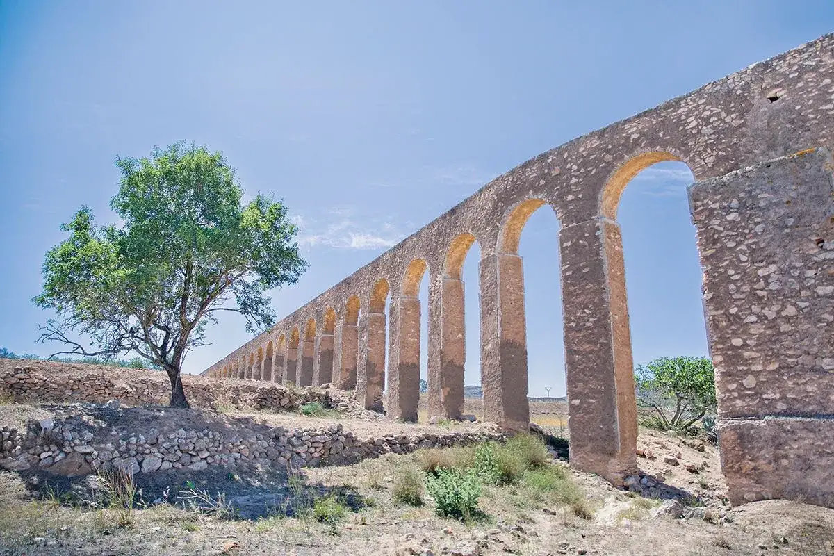 Acueducto romano bajo un cielo azul, con un árbol solitario a la izquierda. Fernán Pérez, explorador de ruinas.