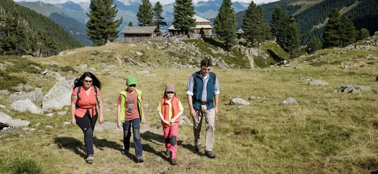 Familie genie&szlig;t eine Bergtour mit Kindern. Sie wandern auf einem grasbewachsenen Hang mit Felsen und B&auml;umen, im Hintergrund Alpenh&auml;user.