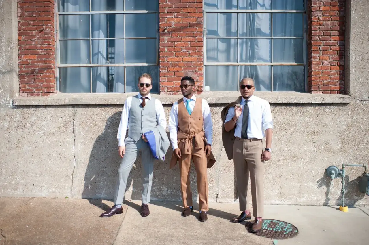 Three men in stylish suits stand against a brick wall, ready for a cocktail attire wedding.