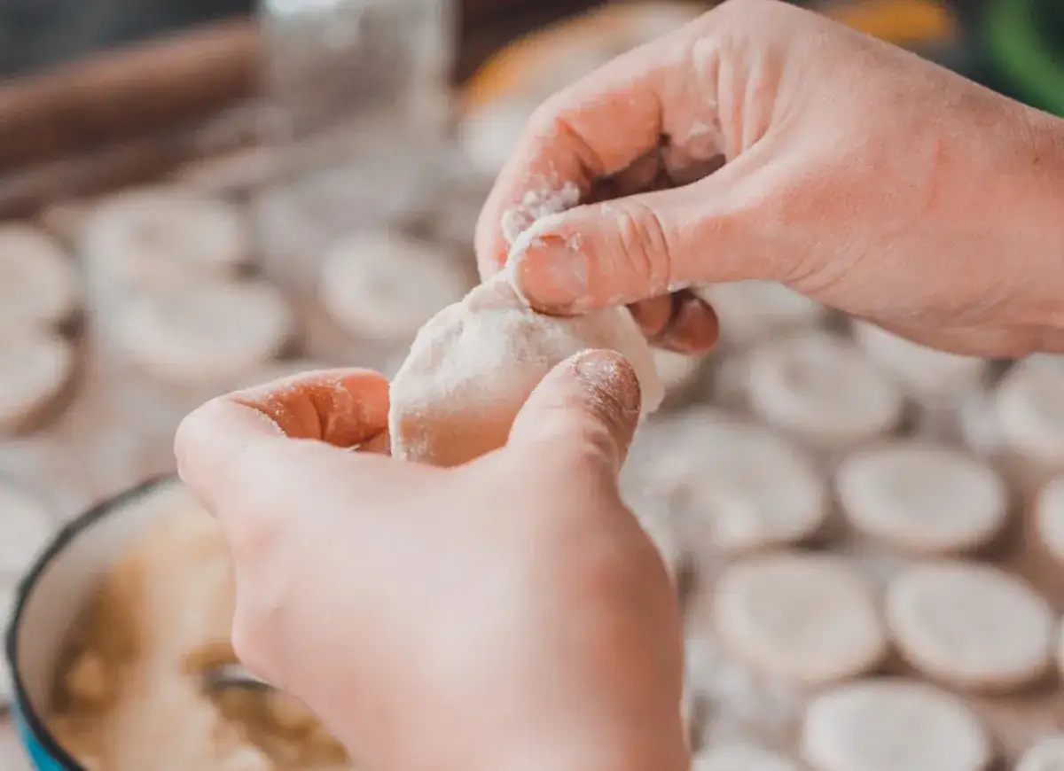 Ręce formują ciasto na pierogi. W tle widać wiele okrągłych kawałków ciasta, gotowych do lepienia.