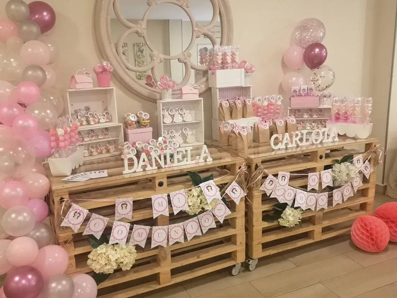 Ideas para mesas de chuches: dos mesas de madera con dulces, globos rosas y blancos, y banderines de comunión.