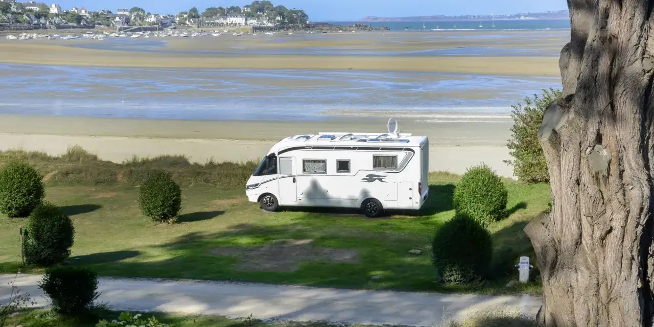 Un camping-car blanc est gar&eacute; sur une pelouse verte, face &agrave; une plage bretonne et un village c&ocirc;tier. Un spot id&eacute;al pour un s&eacute;jour en Bretagne.