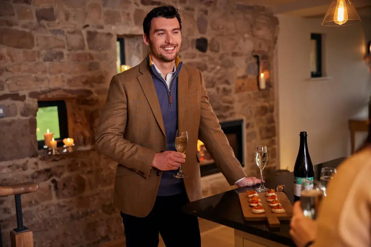 A man in a tweed jacket and blue sweater smiles, holding champagne. Festive chic dress code for a cozy gathering with appetizers and drinks.