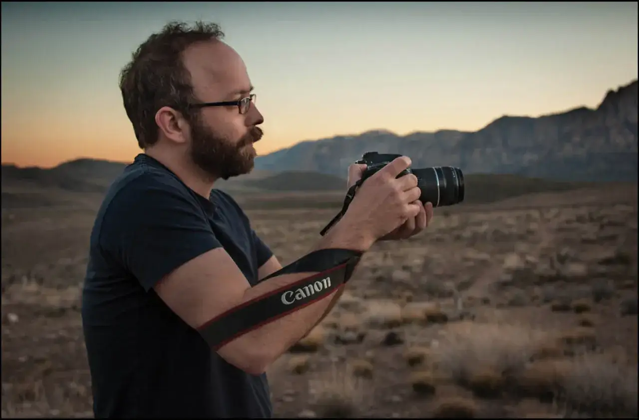 Man with beard and glasses demonstrates how to hold a camera, with a Canon strap around his arm, in a desert landscape at sunset.
