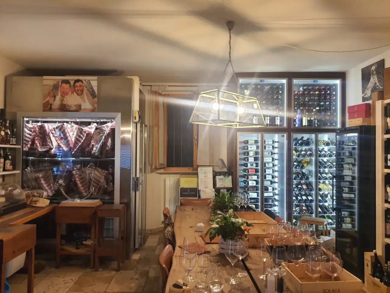Macelleria con carne appesa in vetrina refrigerata, cantine vini illuminate e un lungo tavolo apparecchiato.