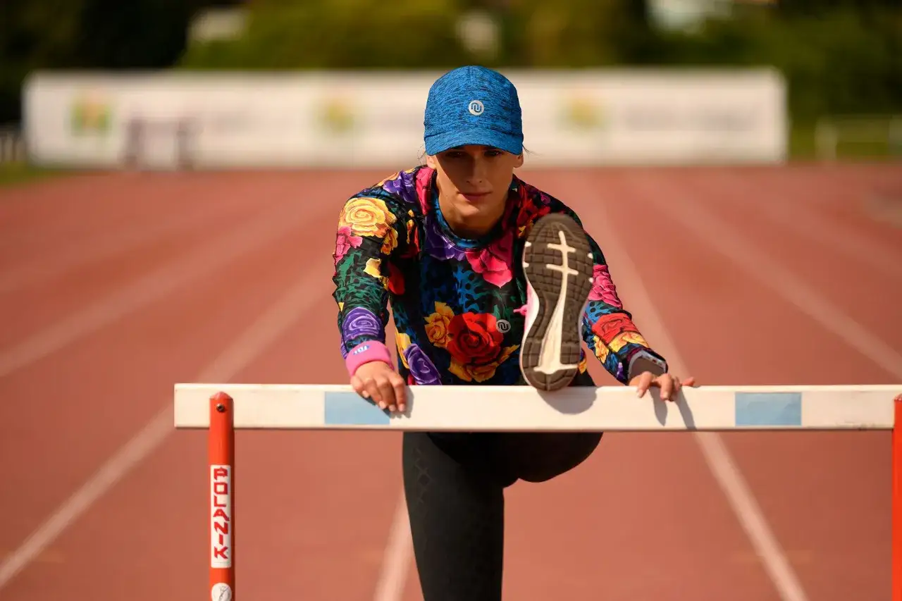 Kobieta w kolorowej bluzie i czapce rozciąga nogę na płotku lekkoatletycznym, przygotowując się do treningu. Pokazuje, jak poprawnie biegać.