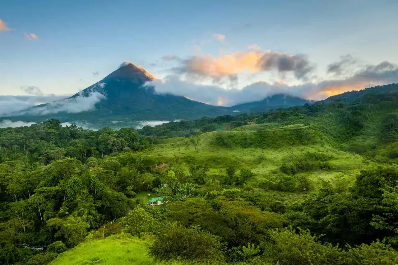 Majestätisk vulkan i Costa Rica reser sig över frodiga, gröna kullar och tät djungel. Moln sveper runt toppen i solnedgångens varma ljus.