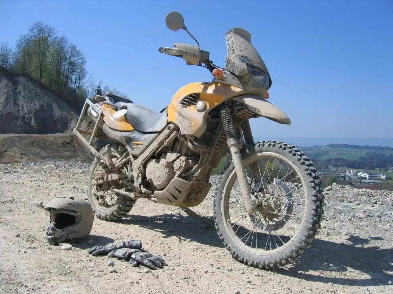 Brudny, żółty motocykl lekkie enduro na kamienistym podłożu, obok leżą kask i rękawice.