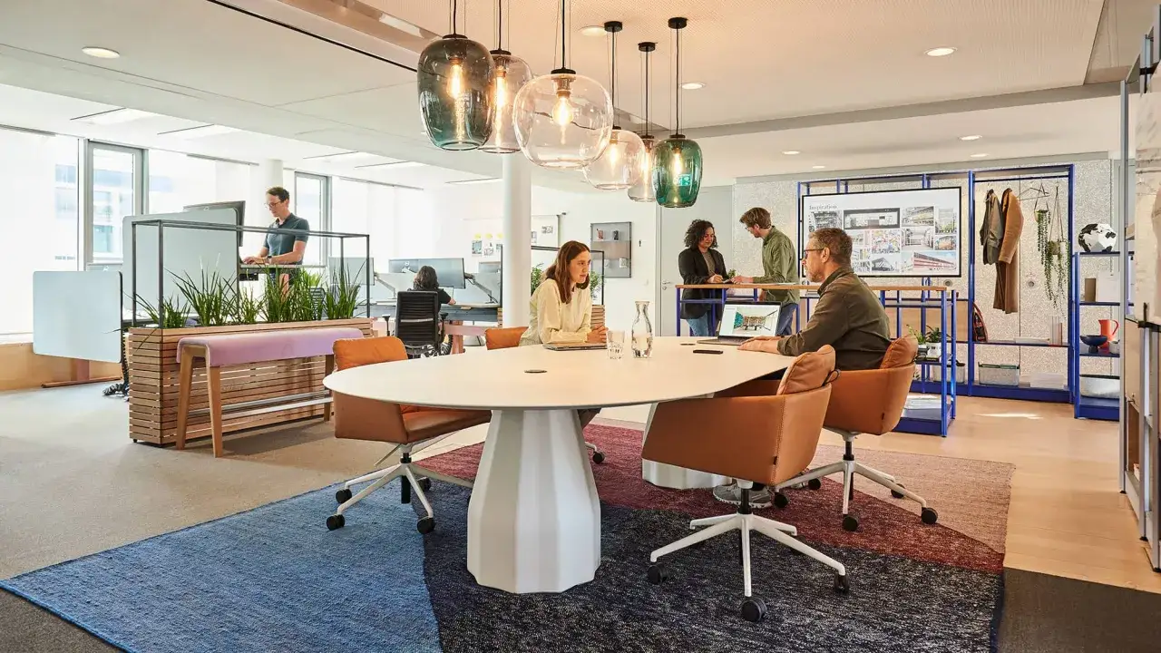 Un espace convivialité entreprise moderne avec une grande table ovale, des fauteuils confortables et des plantes vertes. Des collaborateurs discutent autour de la table.