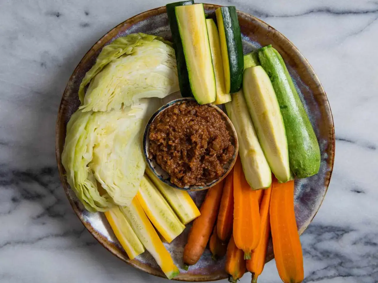 A colorful platter of raw vegetables, including cabbage wedges, zucchini sticks, and carrot sticks, surrounds a small bowl of savory miso dip.