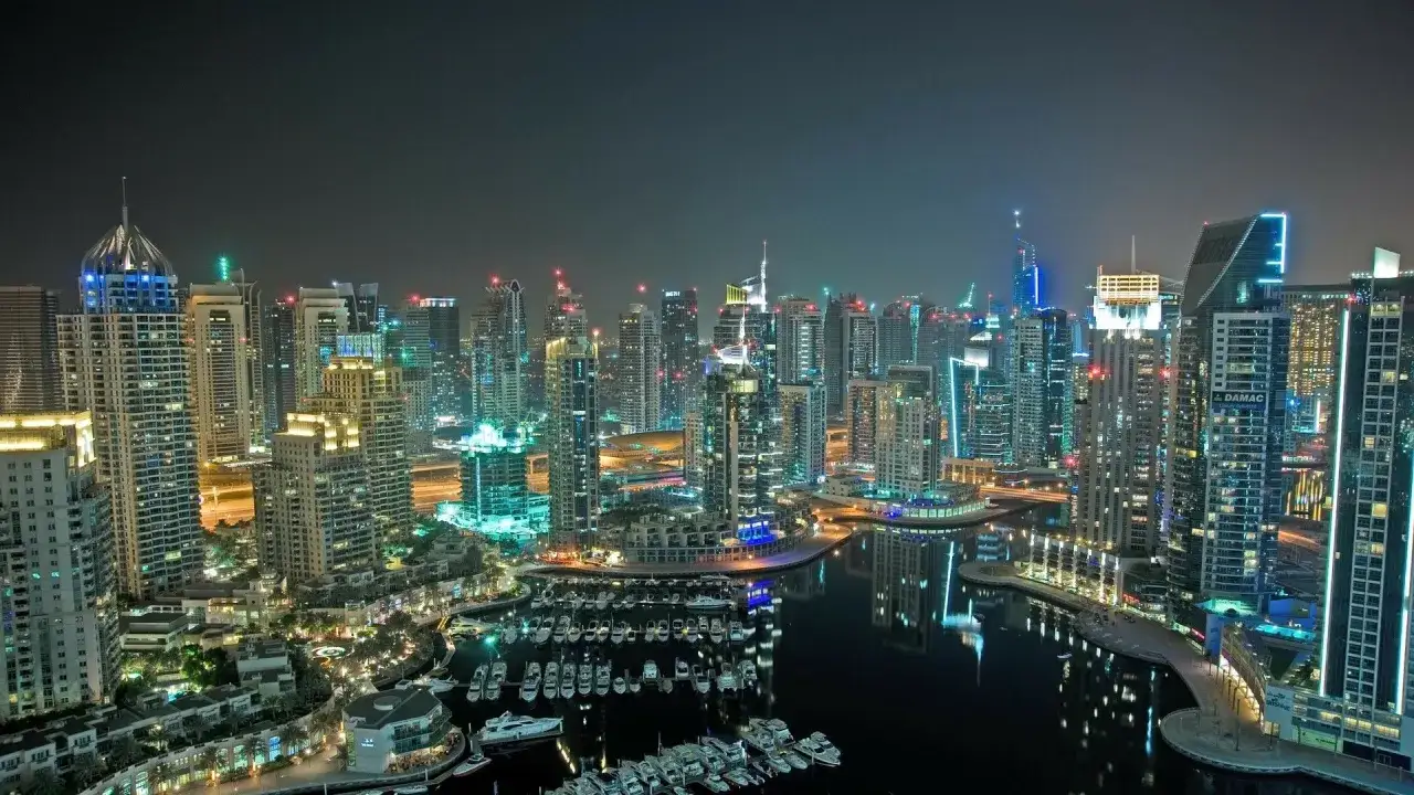 Nocna panorama Dubaju z oświetlonymi drapaczami chmur i mariną pełną jachtów.
