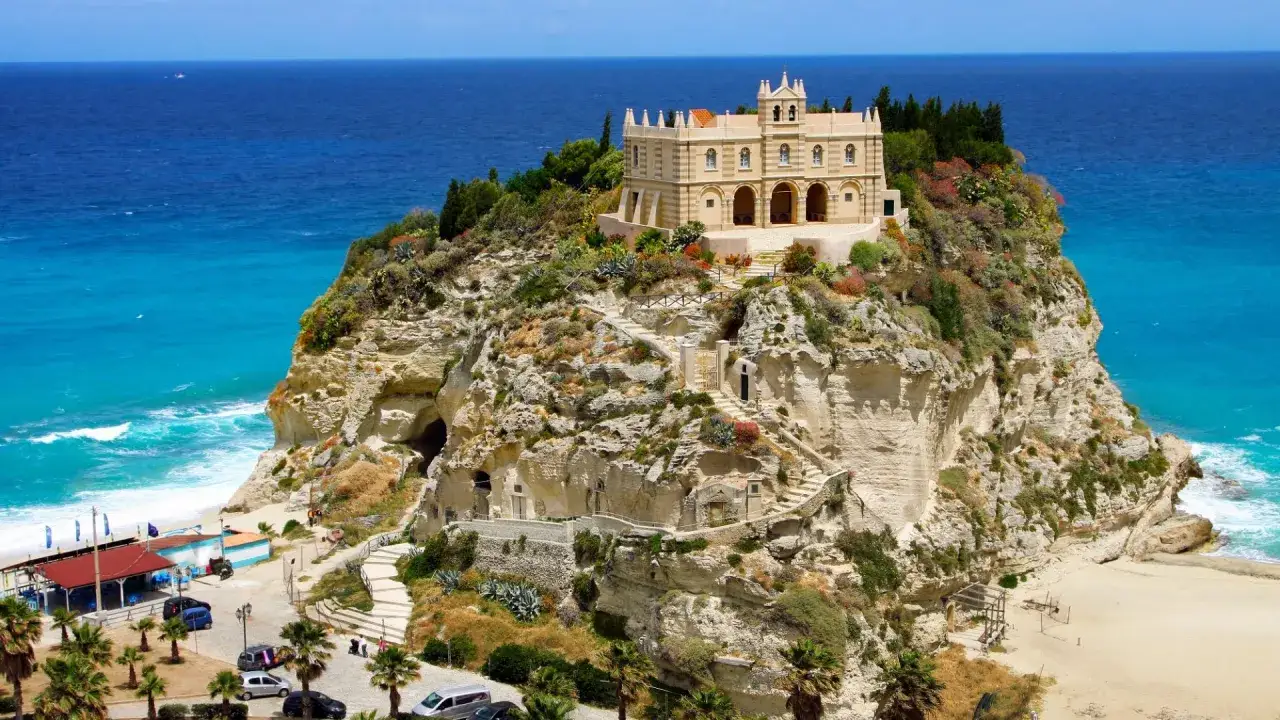 Santuario di Santa Maria dell'Isola su uno sperone roccioso, con il mare in Calabria che lambisce la spiaggia sottostante.