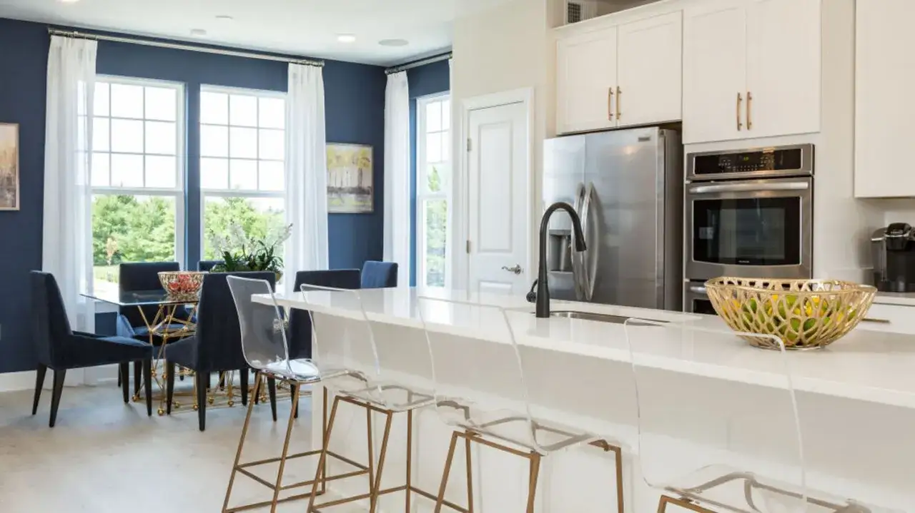 Une cuisine équipée moderne avec un îlot blanc, des tabourets transparents, un réfrigérateur en acier inoxydable et une salle à manger adjacente avec des chaises bleues.
