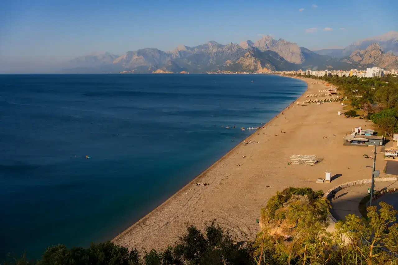 Piękne, szerokie plaże i turkusowe morze w **Antalya miasto**, z g&oacute;rami w tle. Kilka os&oacute;b relaksuje się na piasku, inni pływają.