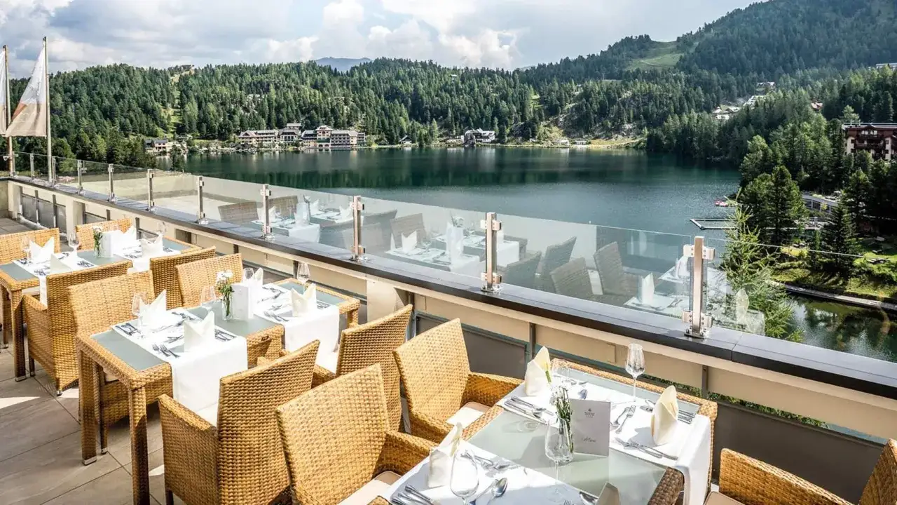 Terrasse mit gedeckten Tischen und Blick auf einen See, umgeben von W&auml;ldern und Hotels auf dem Berg.