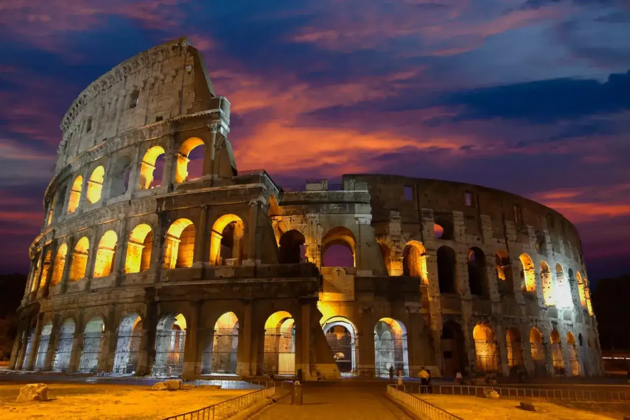 Le Colisée de nuit, illuminé par des lumières chaudes, se dresse sous un ciel aux couleurs flamboyantes. Un spectacle inoubliable.