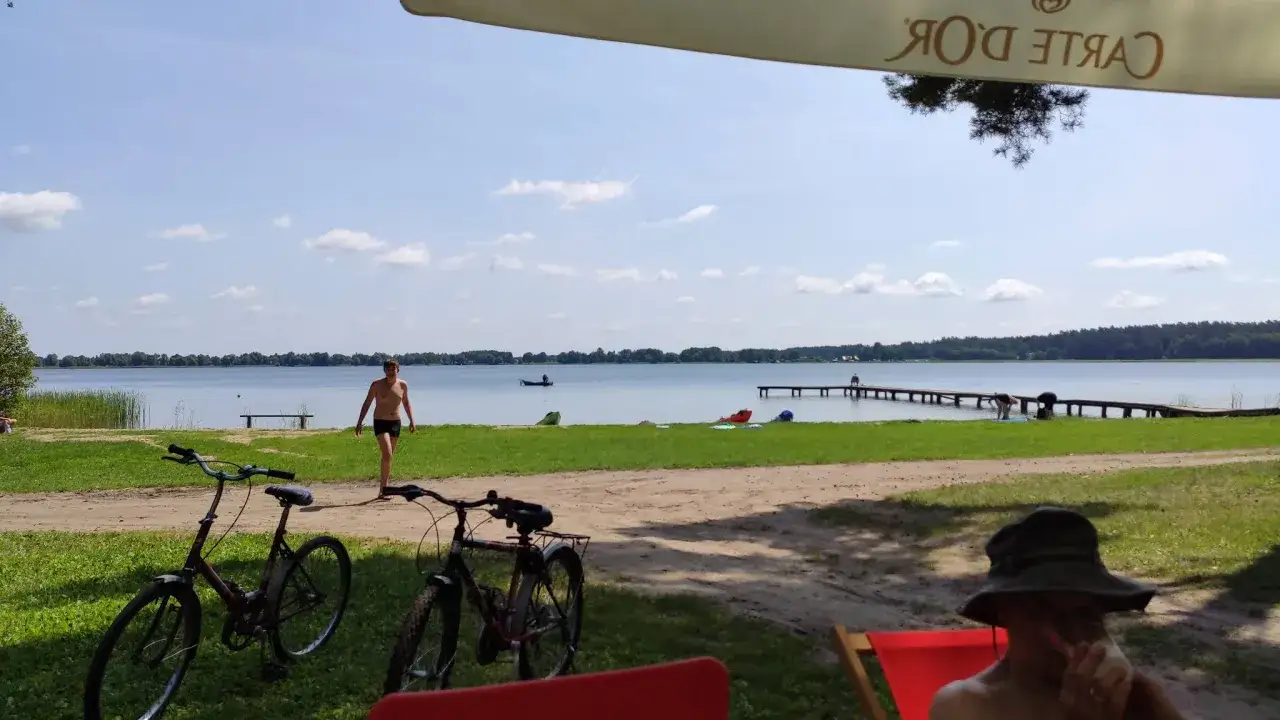 Letnie popołudnie nad jeziorem blisko Warszawy. Dwóch rowerzystów odpoczywa na plaży, podczas gdy inni kąpią się i spacerują po pomoście.