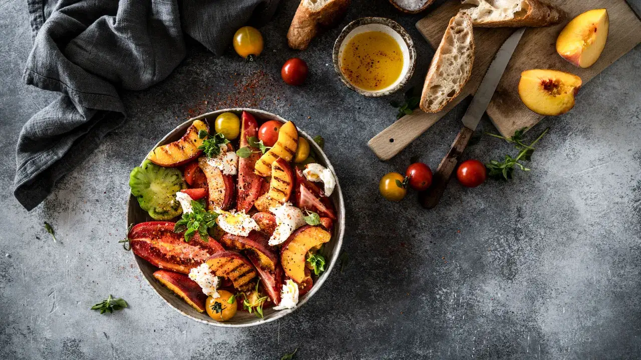 Tomatensalat mit Burrata, gegrillten Pfirsichen und frischen Kr&auml;utern, serviert mit Brot und Oliven&ouml;l.