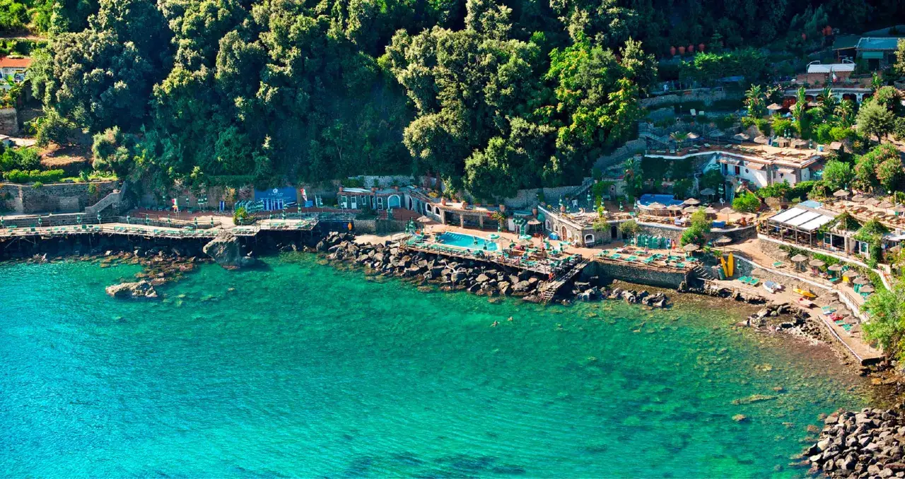 Un incantevole **bagnitiello a Ischia** con acque cristalline, piscine, lettini e ombrelloni su una scogliera verdeggiante.