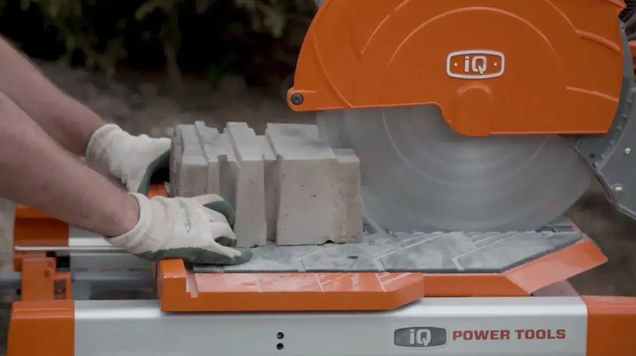 Hands in gloves guide concrete blocks to a spinning blade, demonstrating how to cut brick with an iQ Power Tools saw.