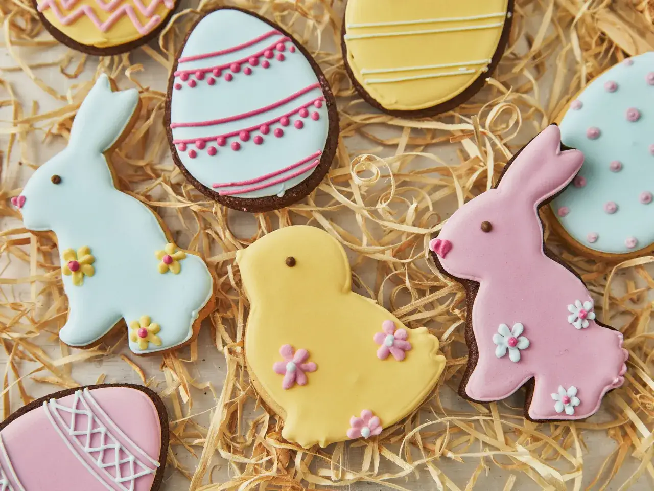 Bunte Osterkekse zum Ausstechen: Hasen, Küken und Eier, verziert mit Zuckerguss und Blumen, liegen auf Heu.