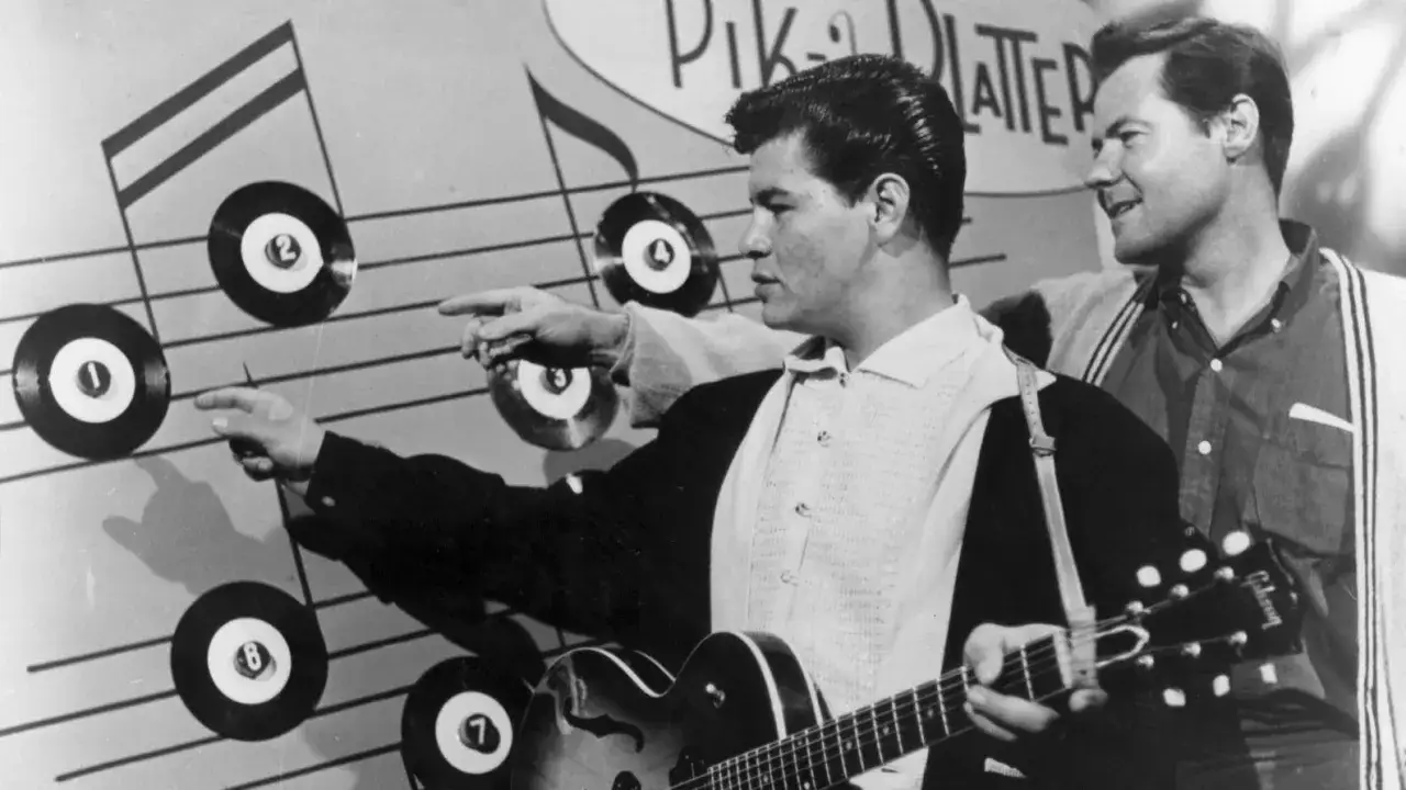 Dwóch muzyków z gitarami na tle nut i płyt winylowych. Jeden z nich wskazuje na płytę z numerem 1, jakby czytał la bamba tekst.