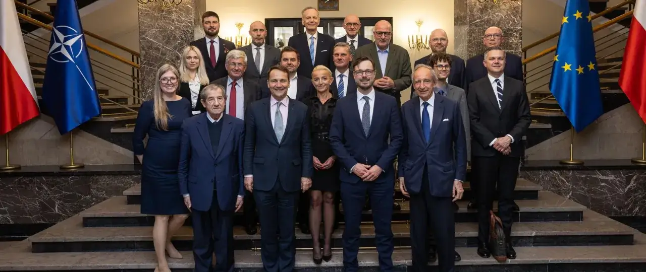 Grupa osób, w tym minister spraw zagranicznych, pozuje do zdjęcia na tle flag Polski, NATO i UE.