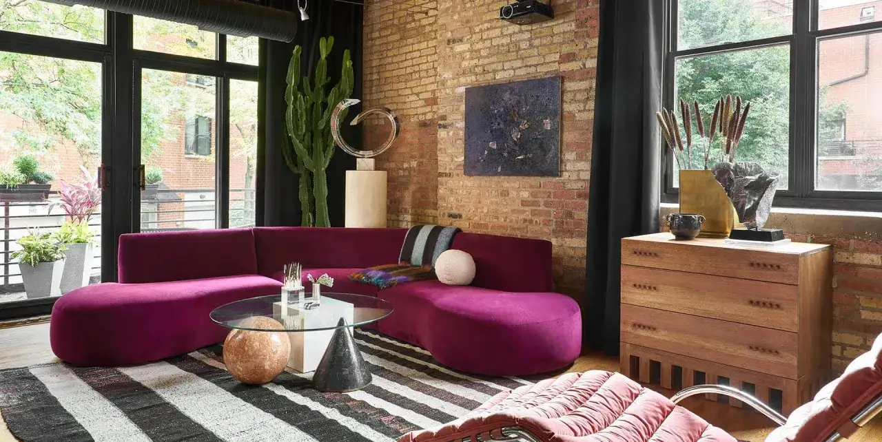 Nowoczesny salon w stylu industrialnym z fioletową sofą, ceglaną ścianą i dużymi oknami.