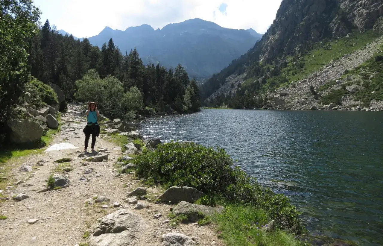 Una mujer camina por un sendero rocoso junto a un estany llong, rodeado de monta&ntilde;as y pinos.