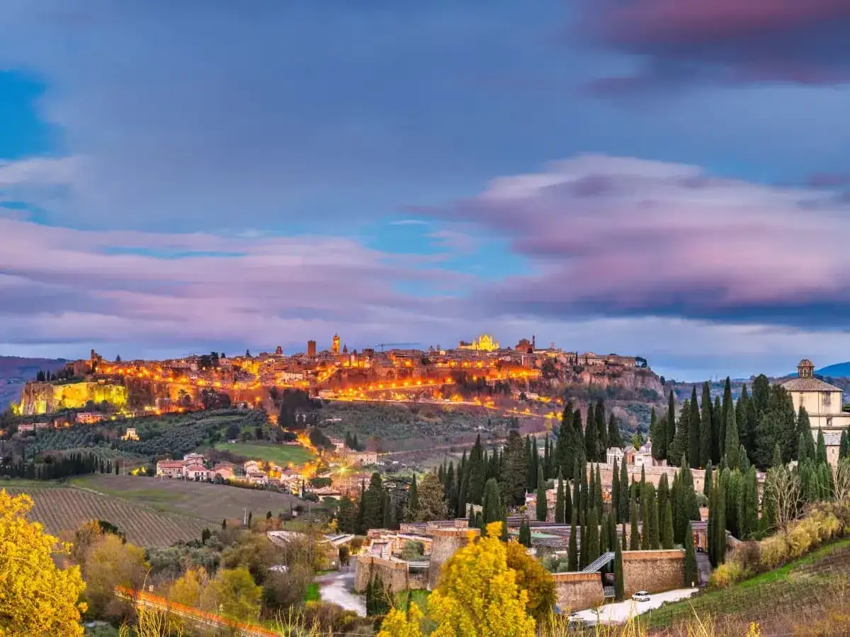 Panoramica di un borgo illuminato su una collina, con cipressi e vigneti in primo piano. Un invito a scoprire i **cammini in Umbria**.