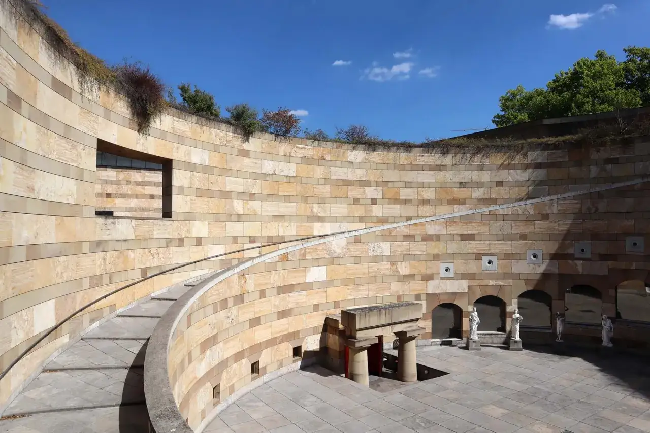 Ein halbrunder Innenhof mit Sandsteinmauern, Treppen und Skulpturen. Die Architektur wirkt **postmodern** und erinnert an ein antikes Amphitheater.