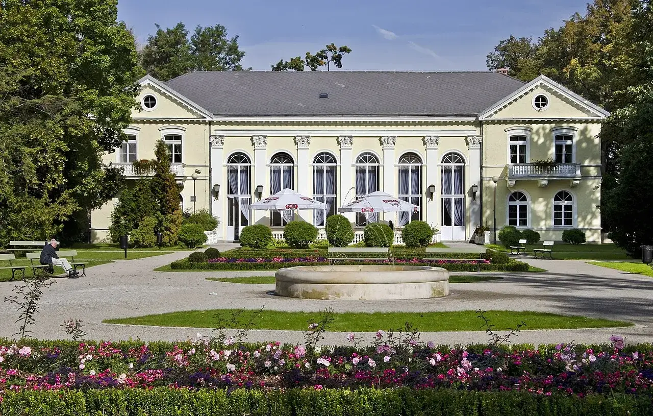 Piękny park w Cieplicach Zdroju z fontanną i kwitnącymi rabatami. W tle zabytkowy budynek, idealne miejsce na relaks i podziwianie atrakcji.