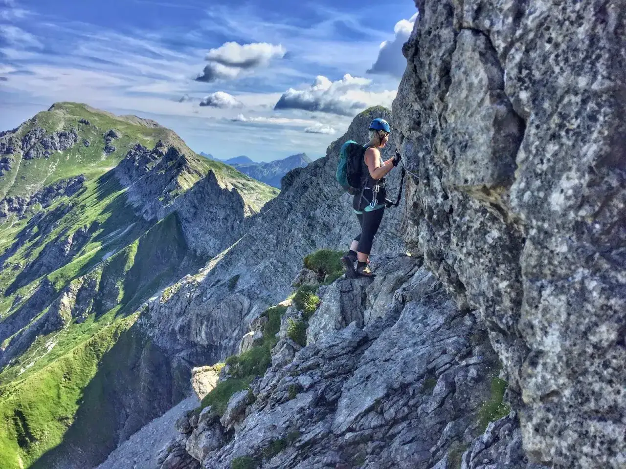 Frau klettert gesichert am Hindelanger Klettersteig mit Blick auf gr&uuml;ne Berge und blauen Himmel.