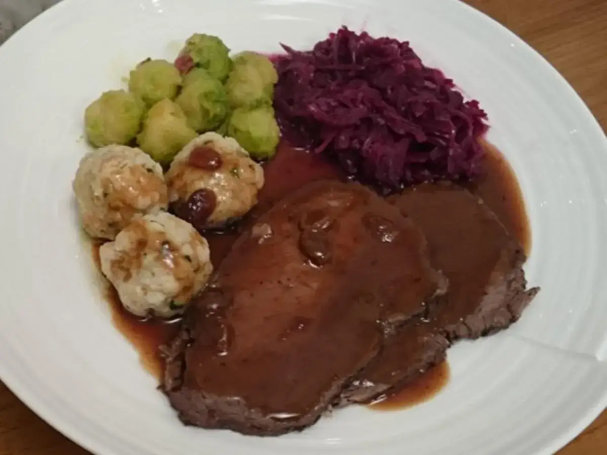 Ein Teller mit Scheiben von Sauerbraten, Kl&ouml;&szlig;en, Rosenkohl und Rotkohl. Ein klassisches Sauerbraten Rezept.