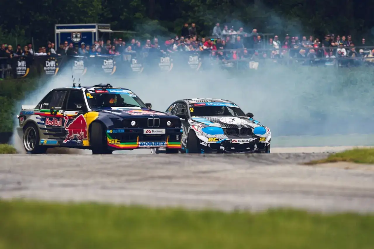 Dwa auta do driftu w akcji, BMW z Red Bull na niebieskim tle i drugie w czarno-biało-niebieskich barwach, generują kłęby dymu na torze wyścigowym.