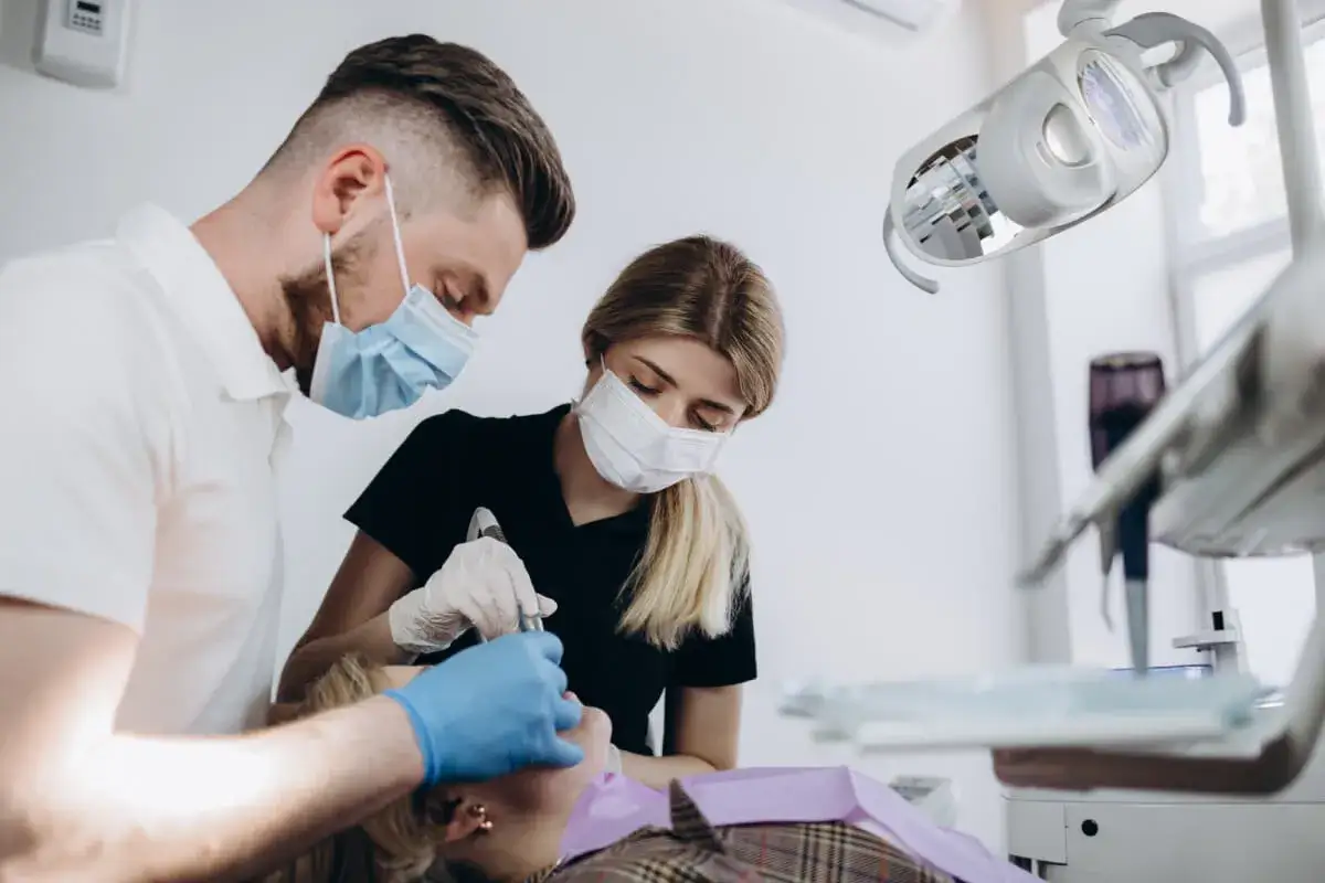 Dentysta i asystentka w maseczkach wykonują zabieg stripping zębów. Pacjentka leży na fotelu.