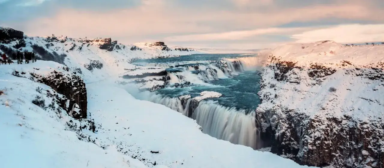 Zimowy krajobraz z potężnym wodospadem Gullfoss. Turkusowa woda spływa kaskadami po ośnieżonych skałach, a na brzegu widać grupki ludzi podziwiających widok.