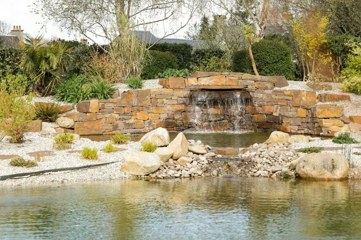 Un magnifique am&eacute;nagement bassin de jardin avec cascade en pierre naturelle, entour&eacute; de v&eacute;g&eacute;tation luxuriante et de galets.