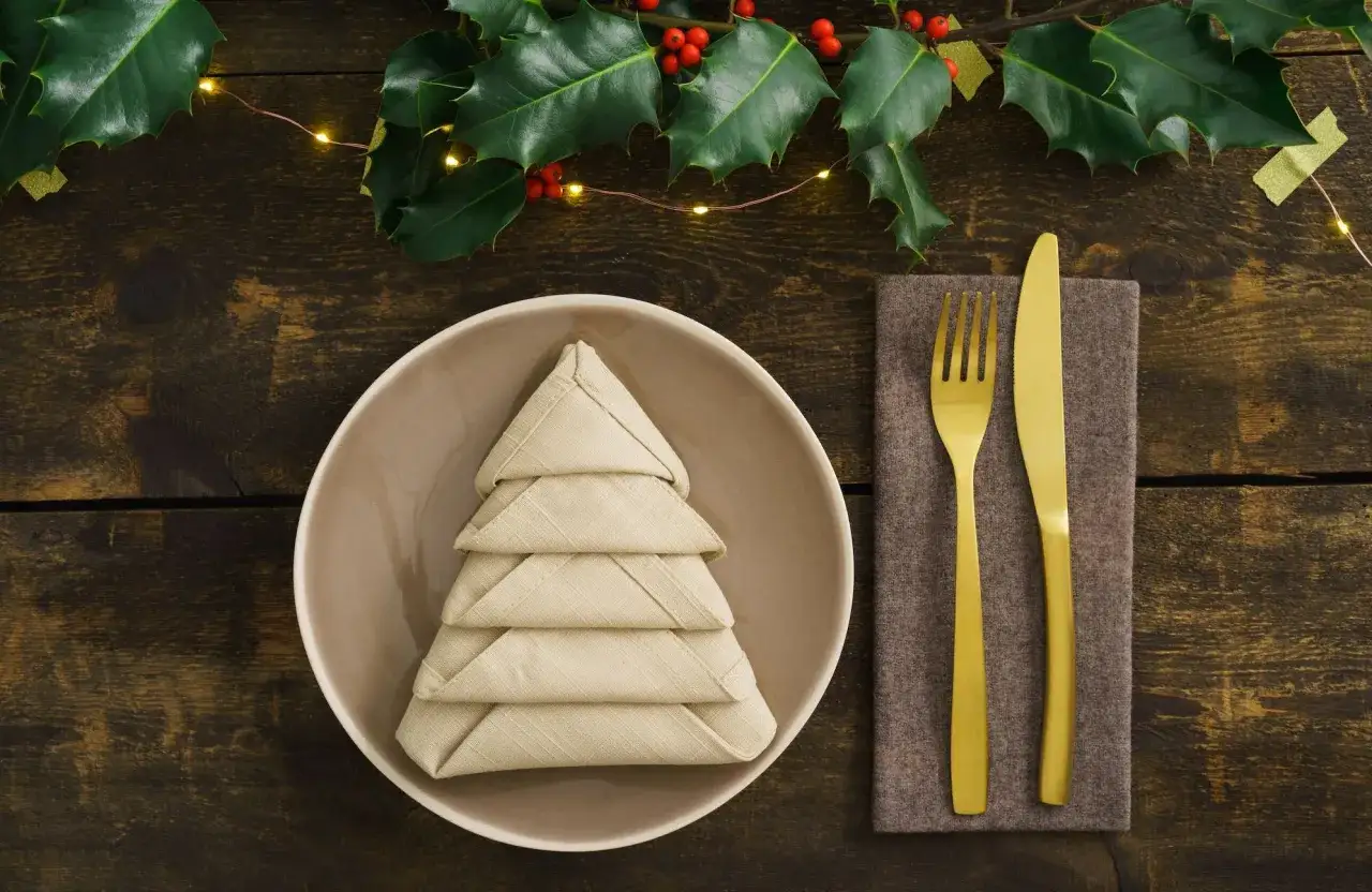 Une assiette avec un pliage serviette tissu noël en forme de sapin, sur une table en bois décorée de houx et de guirlandes lumineuses.