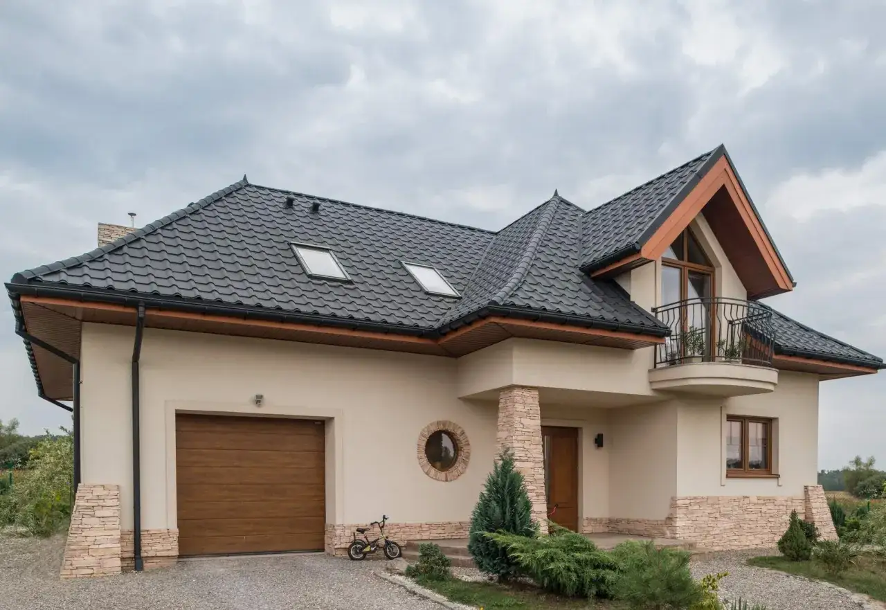 Nowoczesny dom z beżową elewacją, ciemnym dachem i kamiennymi detalami. Widoczny garaż, okrągłe okno i balkon.